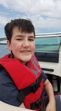 Peter is smiling at the camera. He is sitting in our boat which is on water. He is wearing a red life jacket.