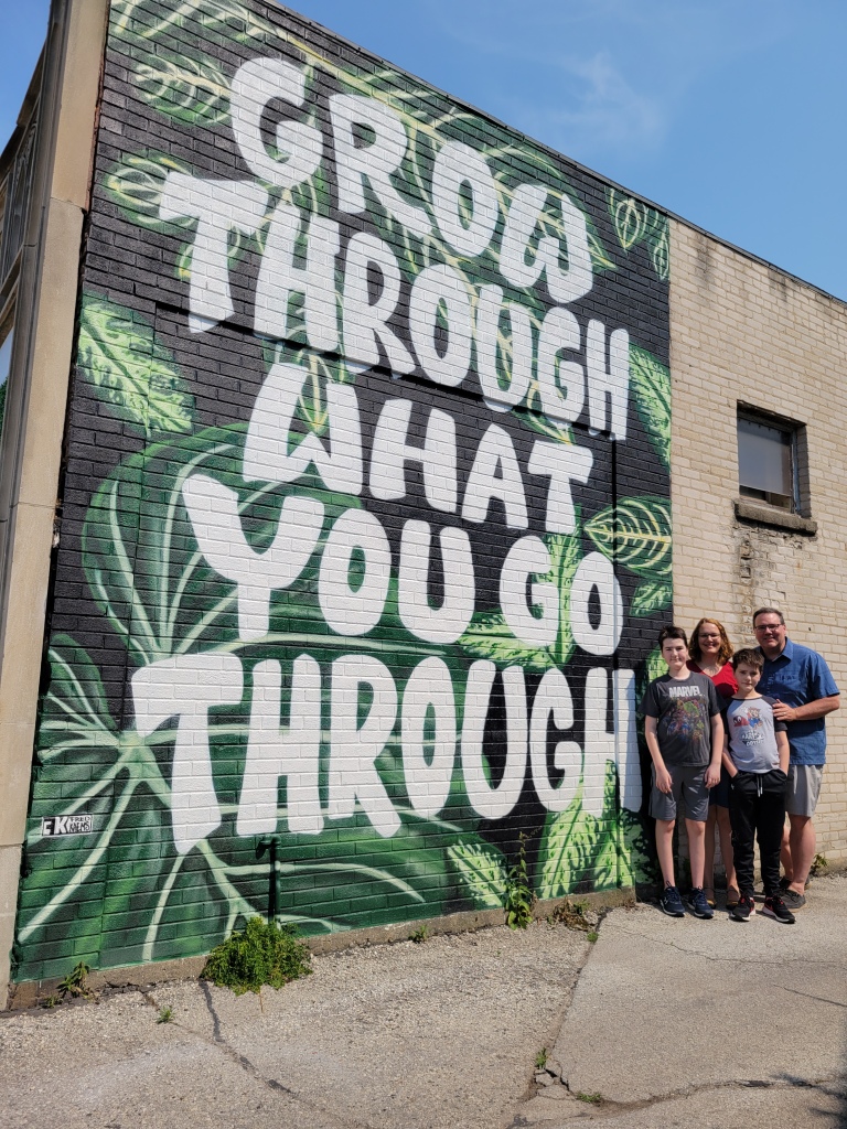 The four of us stand next to a mural painted on the side of a brick building. The background is shades of green and the white lettering says, "Grow Through What You Go Through."